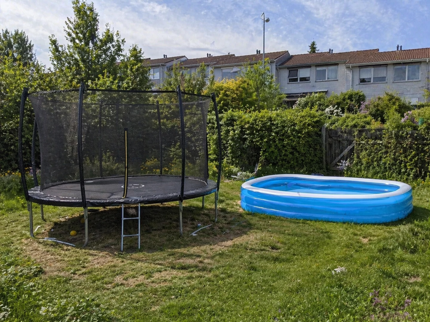 Studsmatta med skyddsnät på gräsyta och en uppblåsbar blå trädgårdspool vid sidan om.